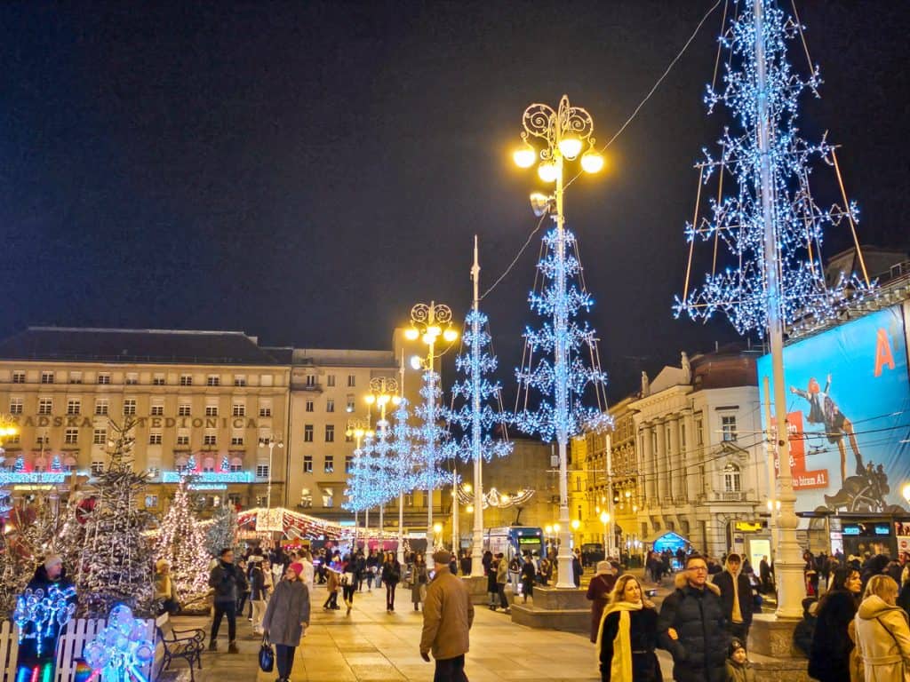 A part of the Zagreb Christmas market is covered with blue lights and street lamps are decorated to look like blue Christmas trees. There are people walking in all directions. 